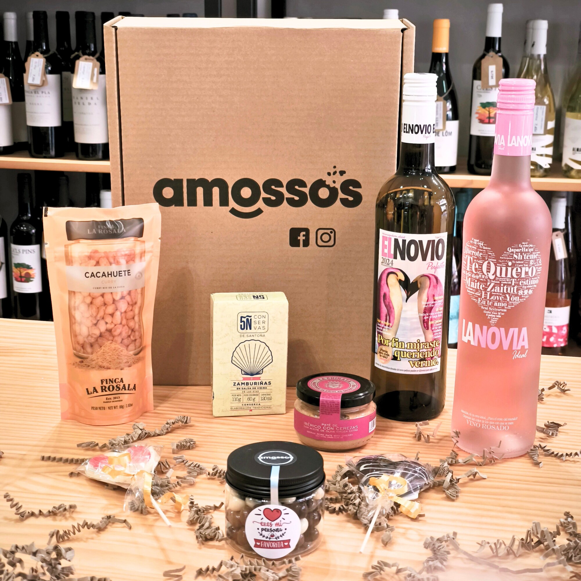 Caja gourmet San Valentín para dos bodegón Pack regalo San Valentín de Amossos con vino El Novio Perfecto, vino La Novia Ideal, zamburiñas, cacahuetes con curry, paté de secreto ibérico con cerezas y dos piruetas de chocolate en forma de corazón sobre mesa.